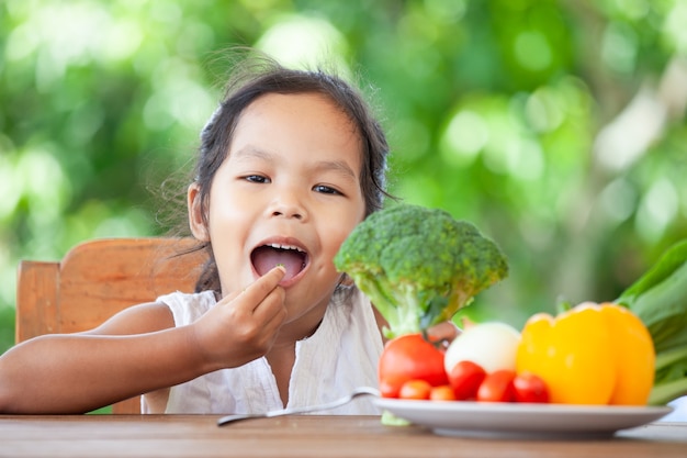 Ragazza asiatica sveglia del bambino che mangia i broccoli e che imparano circa le verdure con felicità