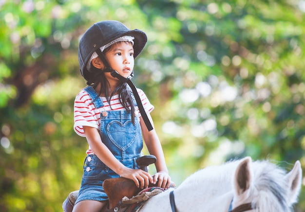 Ragazza asiatica sveglia del bambino che monta un cavallo nell&#39;azienda agricola con divertimento