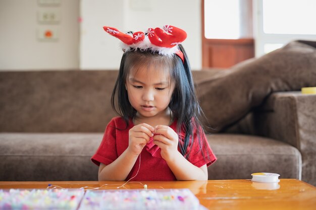 Ragazza asiatica sveglia del bambino che porta il costume di natale che filetta le perle su una stringa con intenzione e divertimento in casa.