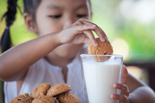 Ragazza asiatica sveglia del piccolo bambino che mangia biscotto con latte per la prima colazione