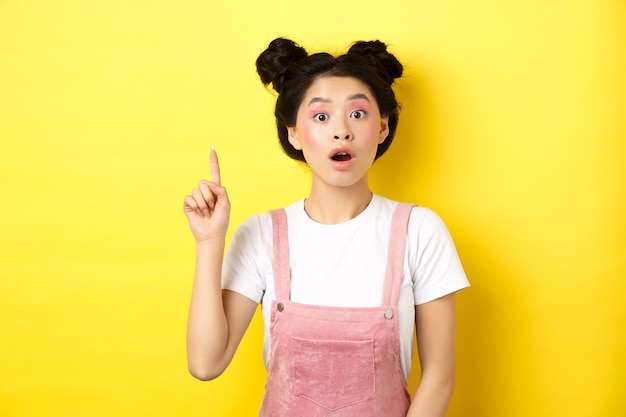 Ragazza asiatica teenager eccitata che ha idea, alzando il dito e dire suggerimento, indossando abiti estivi alla moda con trucco, giallo.