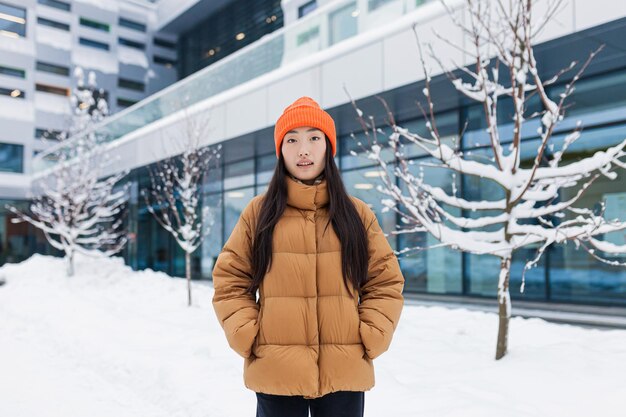 Ragazza asiatica vestita calorosamente che cammina per strada in una giornata nevosa invernale Ritratto di una donna con una giacca e un cappello caldo arancione