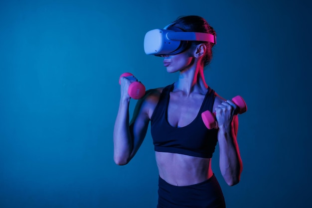 ragazza atletica che fa allenamento online con occhiali VR in abiti neri con manubri su uno sfondo scuro allenamento sportivo online