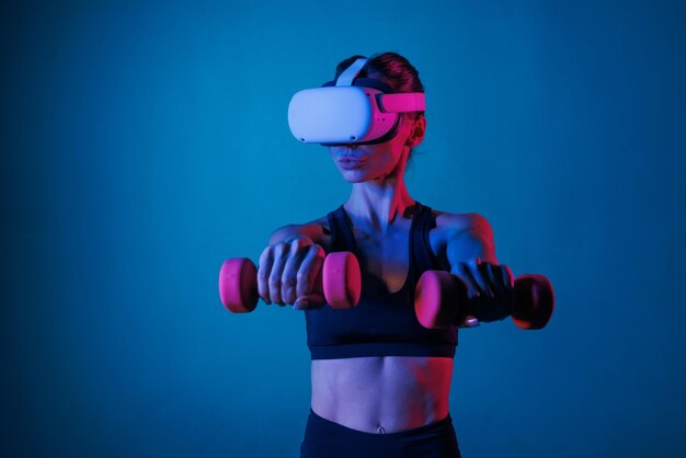 ragazza atletica che fa allenamento online con occhiali VR in abiti neri con manubri su uno sfondo scuro allenamento sportivo online