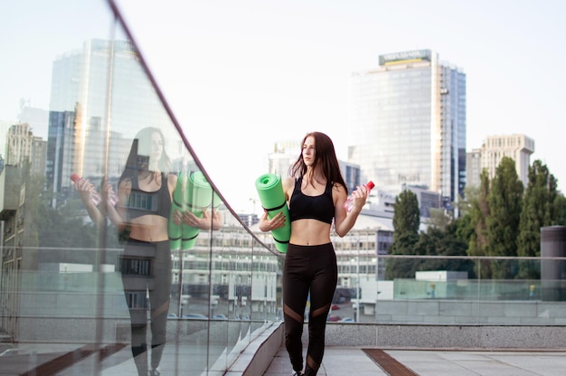 Ragazza atletica che fa yoga al mattino per strada tiene un tappetino sullo sfondo della città