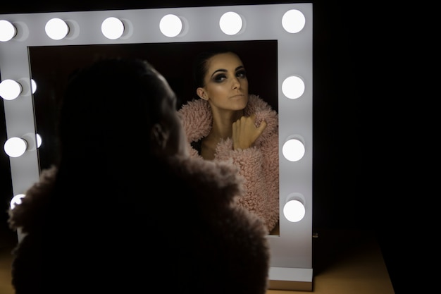 Ragazza attraente che guarda la sua bellezza in uno specchio luminoso con un cappotto rosa