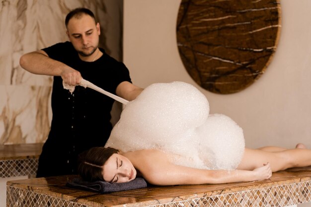 Ragazza attraente che si rilassa nella spa Il massaggiatore sta facendo il peeling con la schiuma nell'hammam termale turco