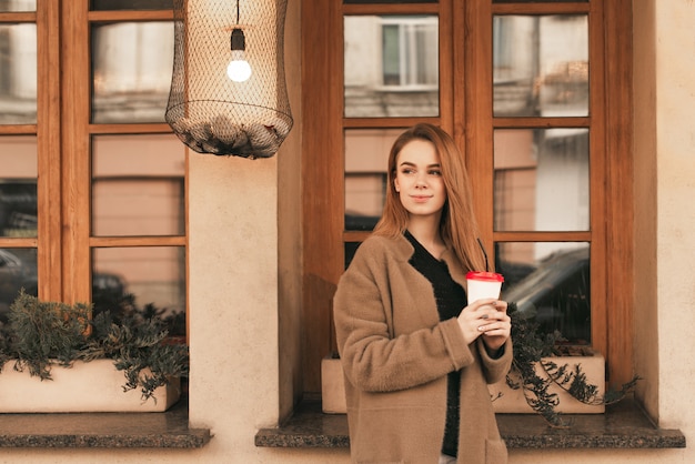 Ragazza attraente con una tazza di caffè in mano, indossa un cappotto beige, in piedi contro il muro con finestre e guardando al lato