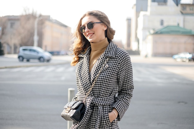 Ragazza attraente in un cappotto in strada in una città, il sole splende, che si rivolge alla telecamera e sorride. ritratto felice rallentatore estate viso tramonto bella signora all'aperto primo piano carino