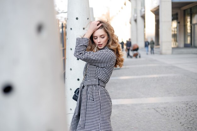 Ragazza attraente in un cappotto in strada in una città, il sole splende, che si rivolge alla telecamera e sorride. ritratto felice rallentatore estate viso tramonto bella signora all'aperto primo piano carino