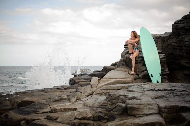 Ragazza attraente nel costume da bagno multicolore seduto vicino alla tavola da surf sulla roccia sopra l'Oceano Atlantico e il cielo limpido