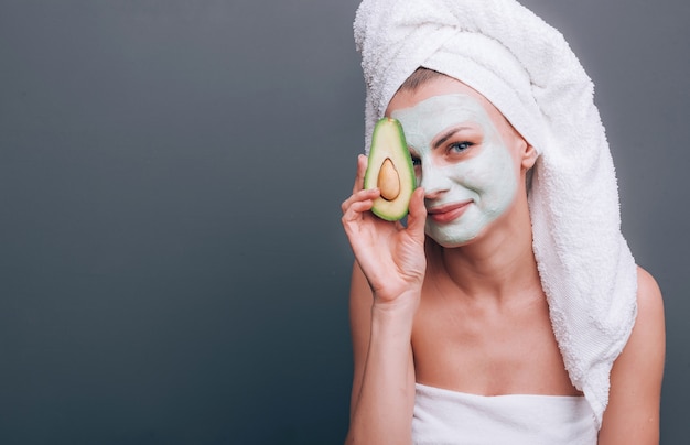 Ragazza avvolta in un asciugamano con una maschera cosmetica sul viso e avocado nelle sue mani