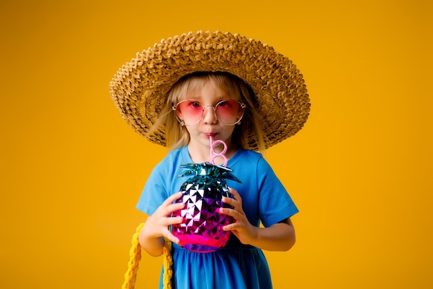 ragazza bambino in un abito blu è in possesso di un bicchiere da bere a forma di ananas su uno sfondo giallo