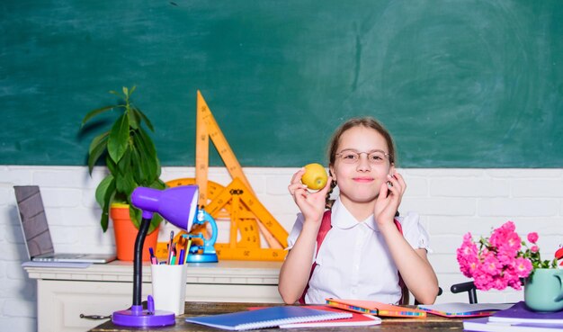 Ragazza bambino piccolo che mangia uno spuntino alla mela Pausa scolastica Rilassati tra le lezioni Carica vitaminica Concetto di vita scolastica Stile di vita sano Studentessa sedersi scrivania sfondo lavagna Kid studente a scuola