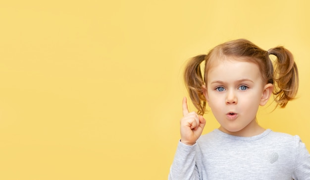 Ragazza bambino sorpreso emozionante che guarda l'obbiettivo e puntare il dito verso l'alto sullo spazio della copia isolato sopra la parete gialla