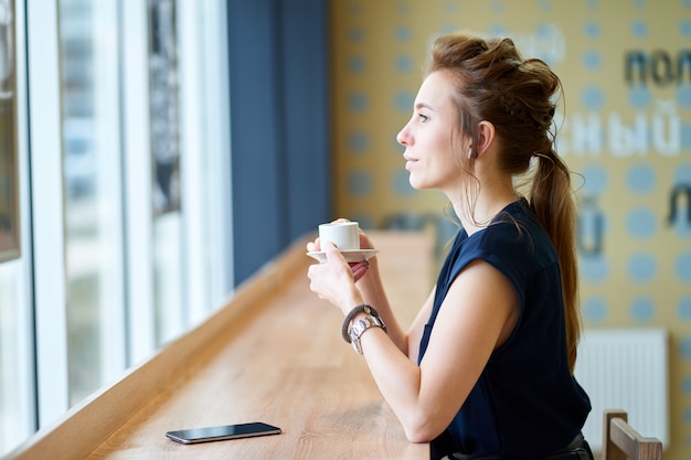 Ragazza bianca rossa in camicia blu scuro, bere il caffè nella caffetteria e guardare alla finestra