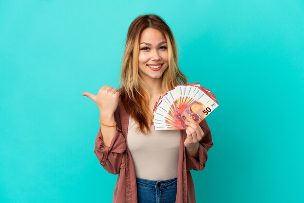 Ragazza bionda adolescente che prende molti euro su sfondo blu isolato che punta al lato per presentare un prodotto present