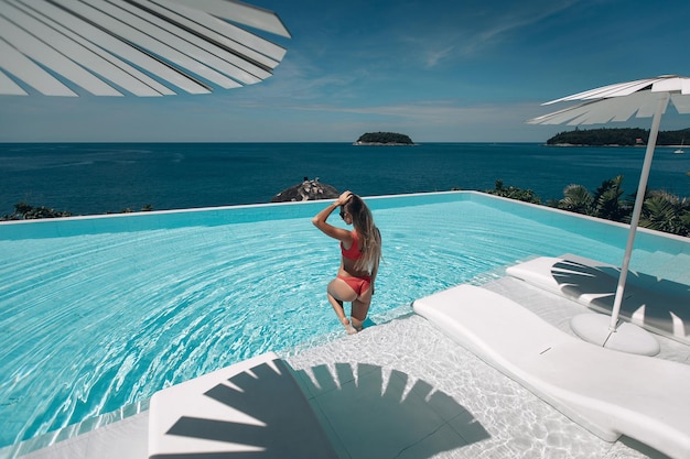 Ragazza bionda affascinante, sexy e calda con un corpo perfetto, in costume da bagno rosso, in posa nella piscina a sfioro. Bellissimo paesaggio marino. Culo sexy. Viaggio a destinazione calda. Phuket. Tailandia. Vista posteriore.