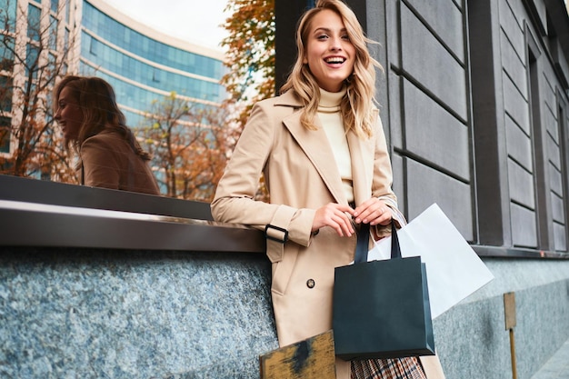 Ragazza bionda alla moda abbastanza allegra in cappotto beige con le borse della spesa che guarda felicemente in macchina fotografica sulla via della città