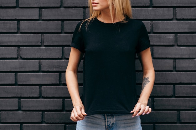 Ragazza bionda alla moda che porta maglietta nera che posa sul fondo nero della parete
