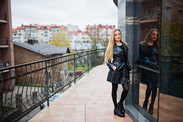 Ragazza bionda alla moda con un lungo cappotto di pelle nera posata contro la grande finestra dell'edificio