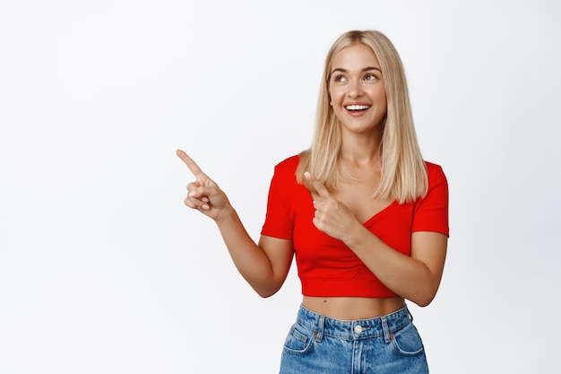 Ragazza bionda allegra sorride punti e guarda da parte al logo aziendale banner mostra pubblicità sullo spazio di copia sinistra si erge su sfondo bianco