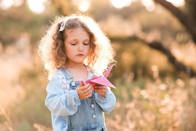 Ragazza bionda carina che tiene in mano una gru di origami di carta in piedi all'aperto