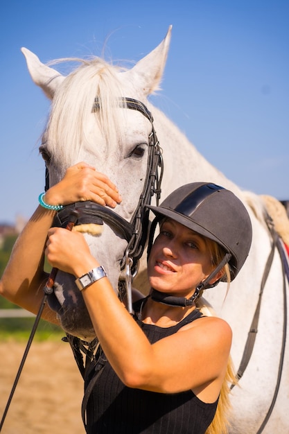 Ragazza bionda caucasica che abbraccia la testa di un cavallo bianco a cavallo, vestita di cavaliere nero con cappello di sicurezza
