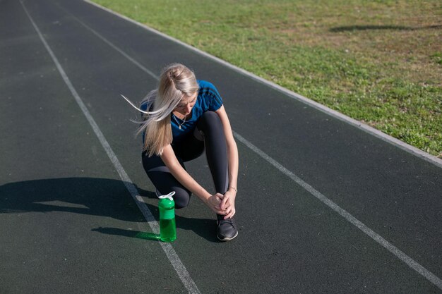 Ragazza bionda che ha infortunio alla caviglia in pista da corsa. Sanità e concetto di sport. bottiglia sportiva verde per bere