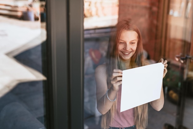 Ragazza bionda che mostra un segno di carta bianca attraverso la finestra di vetro, la nuova normalità