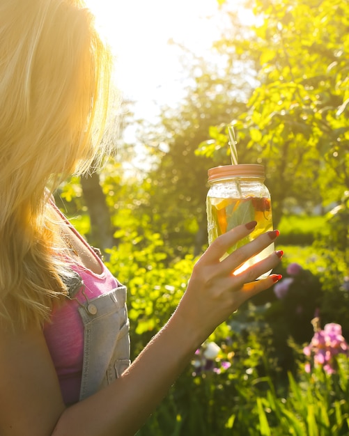 Ragazza bionda che tiene limonata fresca in barattolo con paglia. Bevande estive hipster. Stile di vita vegano sano. Ecologico nella natura. Limoni, arance e frutti di bosco con menta nel bicchiere.