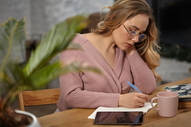 Ragazza bionda con gli occhiali, cardigan lavorato a maglia rosa tiene in mano una penna blu, seduto al tavolo di legno con un tablet, un taccuino, una tazza e un fiore in vaso su di esso. Studente, blogger. Concetto di lavoro o istruzione. Avvicinamento