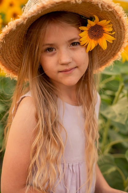 ragazza bionda con i capelli lunghi in un cappello di paglia e un abito di lino rosa si trova in un campo con girasoli