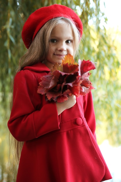 RAGAZZA BIONDA CON I CAPELLI LUNGHI IN UN CAPPOTTO ROSSO E UN BERRETTO ROSSO È IN PIEDI DA UN SALICE AUTUNNO