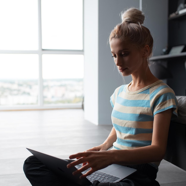 Ragazza bionda con il panino per capelli che lavora a casa digitando sul computer portatile seduto sul pavimento