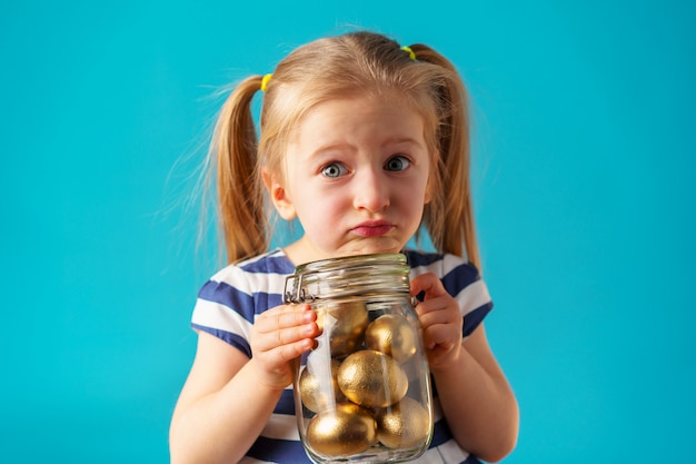 Ragazza bionda del bambino con il barattolo di vetro pieno di uova dipinte dorate per Pasqua