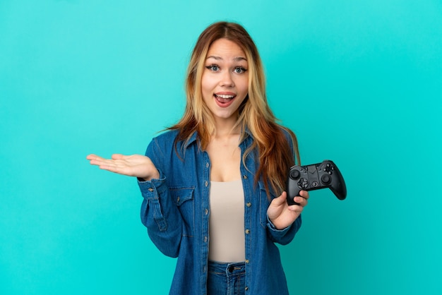 Ragazza bionda dell'adolescente che gioca con un controller per videogiochi su un muro isolato con espressione facciale scioccata