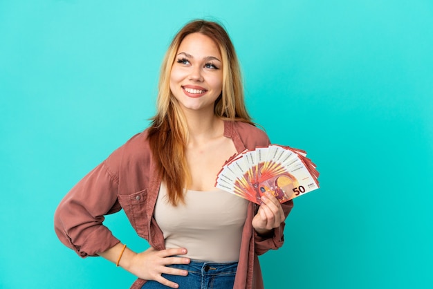 Ragazza bionda dell'adolescente che prende molti euro sopra fondo blu isolato che posa con le armi all'anca e che sorride