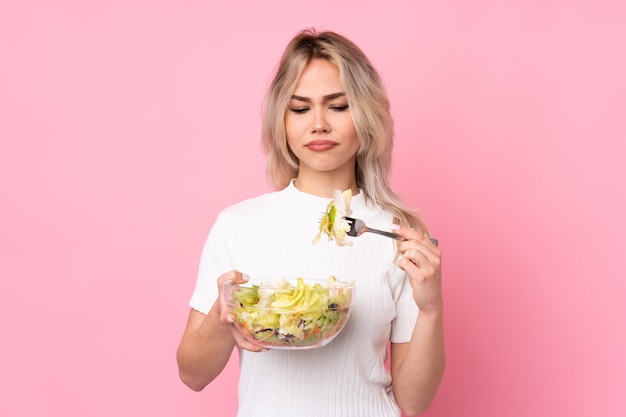 Ragazza bionda dell'adolescente che tiene un'insalata sopra la parete rosa isolata