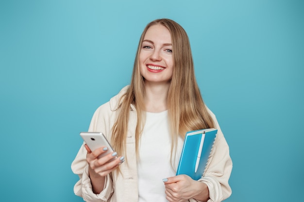 Ragazza bionda dell'allievo che tiene telefono cellulare e blocco note e sorridente isolato sulla parete blu. Formazione in linea