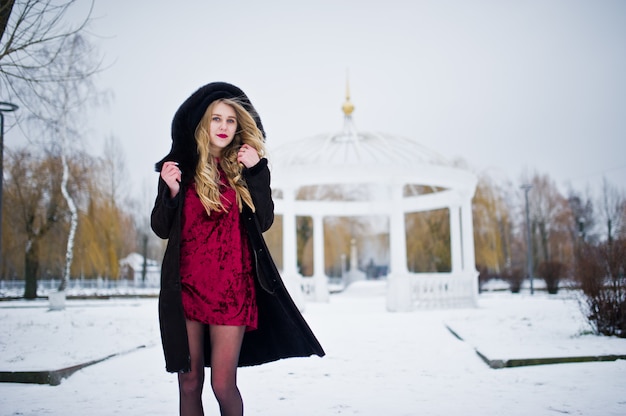 Ragazza bionda di eleganza in pelliccia e vestito da sera rosso che posano al giorno nevoso di inverno.