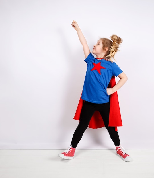 Ragazza bionda di sei anni vestita da supereroe che si diverte a casa. Muro bianco su sfondo.