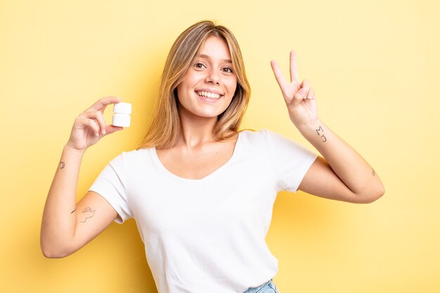 Ragazza bionda graziosa che sorride e che sembra amichevole, mostrando il numero due. concetto di bottiglia di pillole