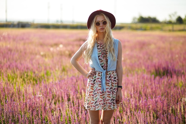 Ragazza bionda hipster in cappello in posa su un campo floreale isolato sfondo tramonto