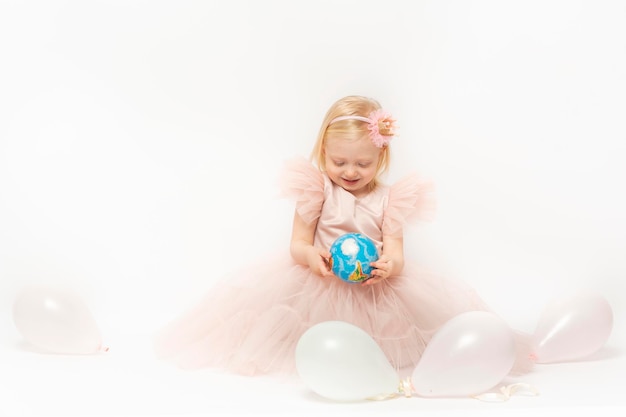 Ragazza bionda in abito rosa si siede su sfondo bianco con palloncini Il bambino ride e guarda il piccolo globo nelle mani