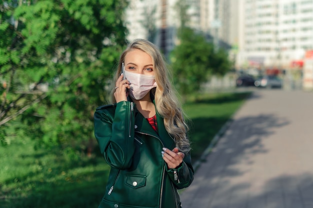 Ragazza bionda in maschera medica bianca e giacca verde che parla sul telefono