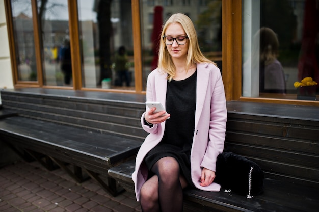 Ragazza bionda in occhiali e cappotto rosa, tunica nera seduto sulla panchina in strada e guardando su smartphone.