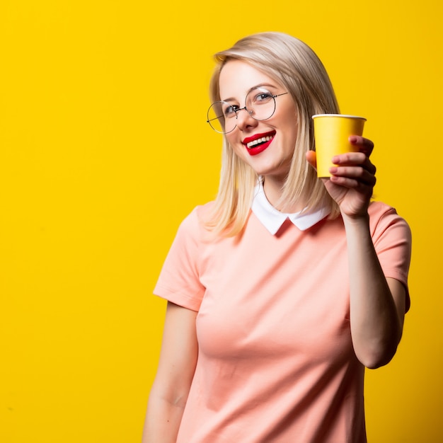 Ragazza bionda in vestito rosa con la tazza di carta su spazio giallo