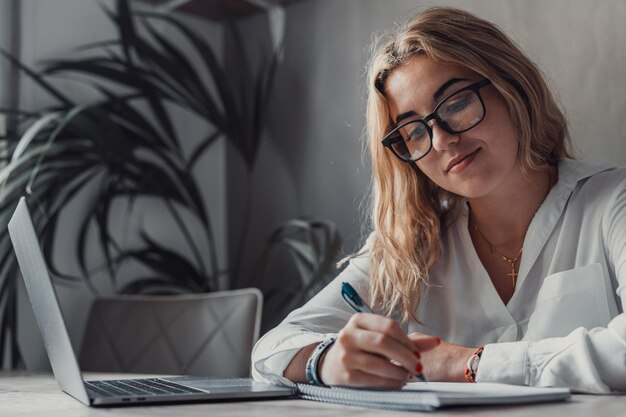 Ragazza bionda millenaria seduta alla scrivania nel salotto che studia sul portatile che prende appunti concentrata giovane donna che lavora al computer che scrive in un quaderno prende un corso online o una formazione a casa concetto di istruzione xA