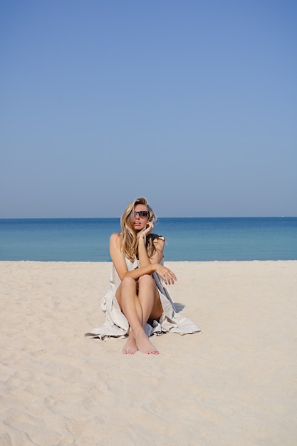Ragazza bionda sorridente che si siede alla spiaggia sabbiosa in una mattina d'estate. Outdoor ritratto di bella donna sorridente sul mare blu e sullo sfondo del cielo sereno.